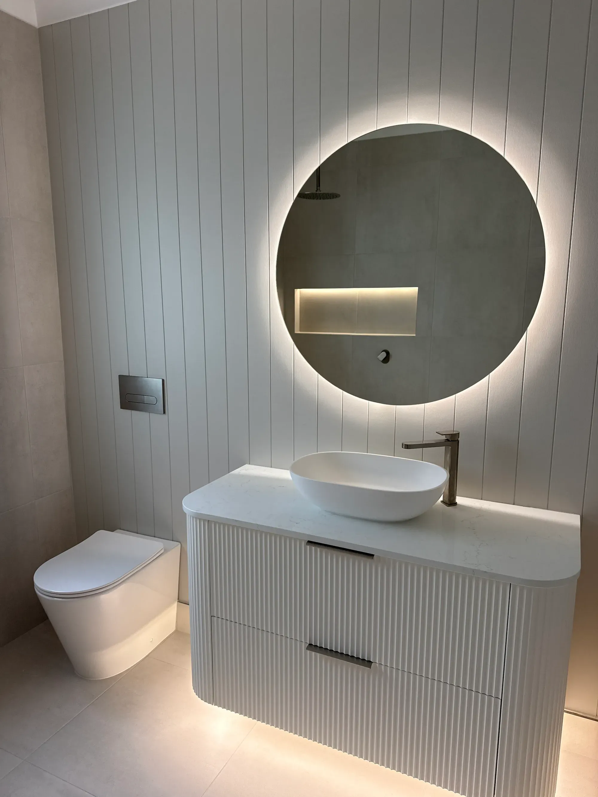 Fluted wall-hung vanity with white vessel basin, brushed nickel mixer and a back-lit round mirror in a completed bathroom
